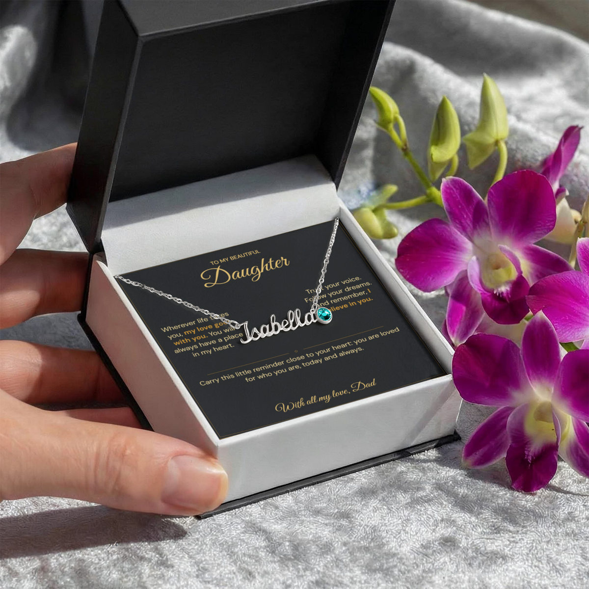 Hand holding an open black jewellery box with an Isabella name necklace, blue birthstone charm and Daughter message card beside purple flowers.