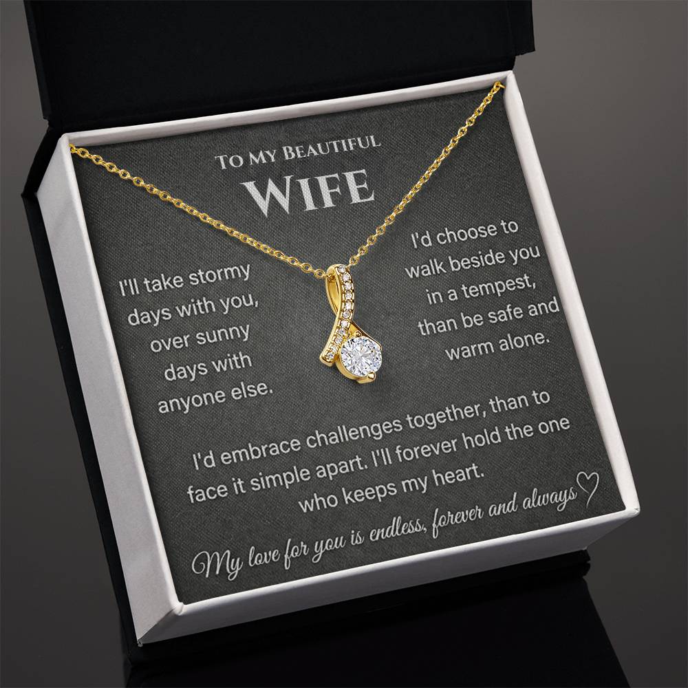 Angled view of a gold-toned ribbon-shaped necklace on a wife message card in an open box.
