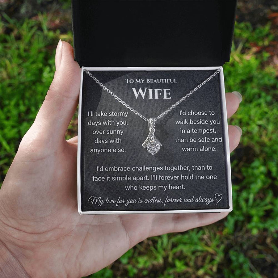 A hand holds an open jewellery box containing a silver-toned necklace and wife message card outdoors.