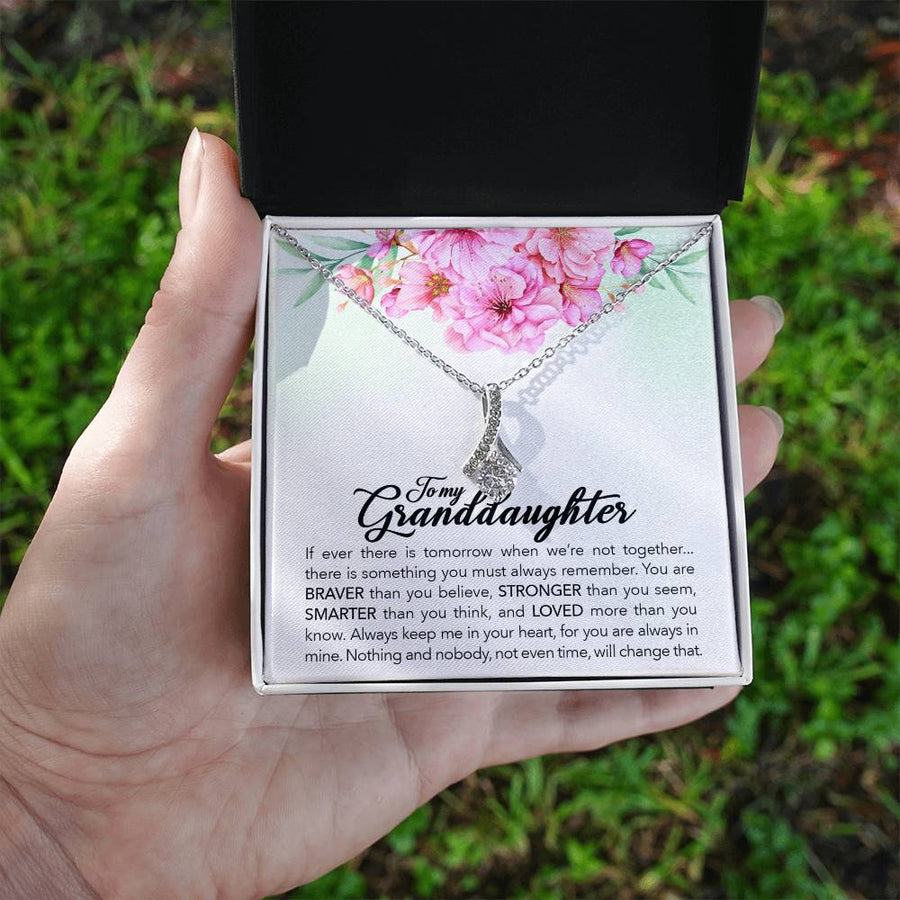 Hand holding an open black jewellery box with a silver-tone ribbon pendant and floral granddaughter message card outdoors.