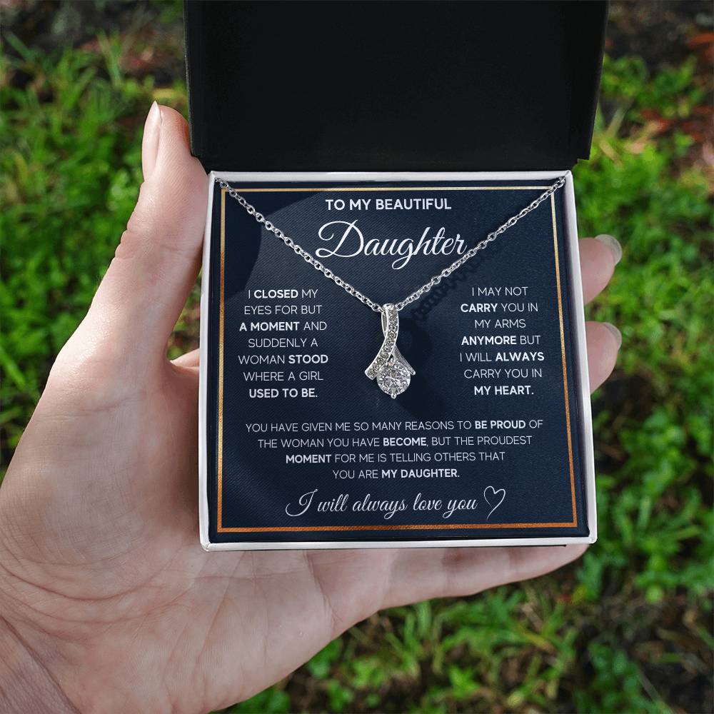 A hand holds an open white jewellery box showing a silver ribbon pendant and daughter message card