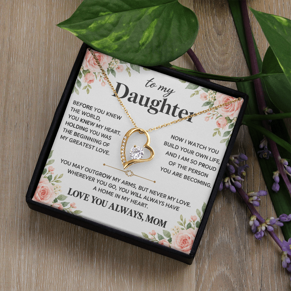 Gold-tone heart pendant necklace in an open black box surrounded by green leaves and flowers.